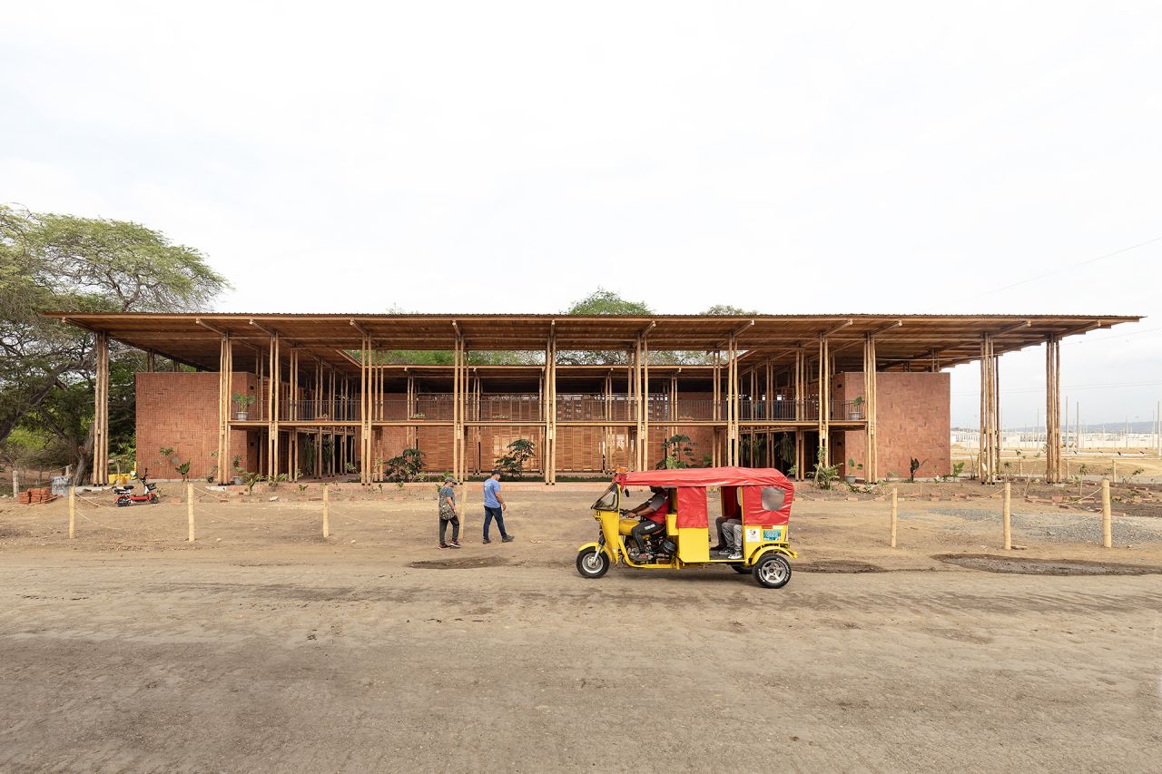 A large building is being constructed. Two people walk toward it.  a yellow and red auto rickshaw is parked in the foreground on a dirt road. The surrounding landscape is arid, with sparse vegetation.