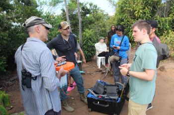 Members of the Welcome to the Jungle team do fieldwork outside. In the foreground, three men are talking with one holding an orange device. A woman is using a mobile device near them. Other people are sitting or standing in the background. There is a black container with scientific equipment on the ground. The background is filled with trees.